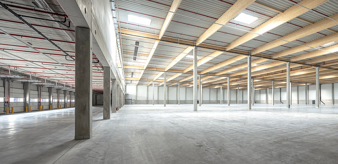 Weitläufiger, heller Lagerraum mit hohen Decken und Betonstützen, großen Fensteröffnungen und einem klaren Fußboden.