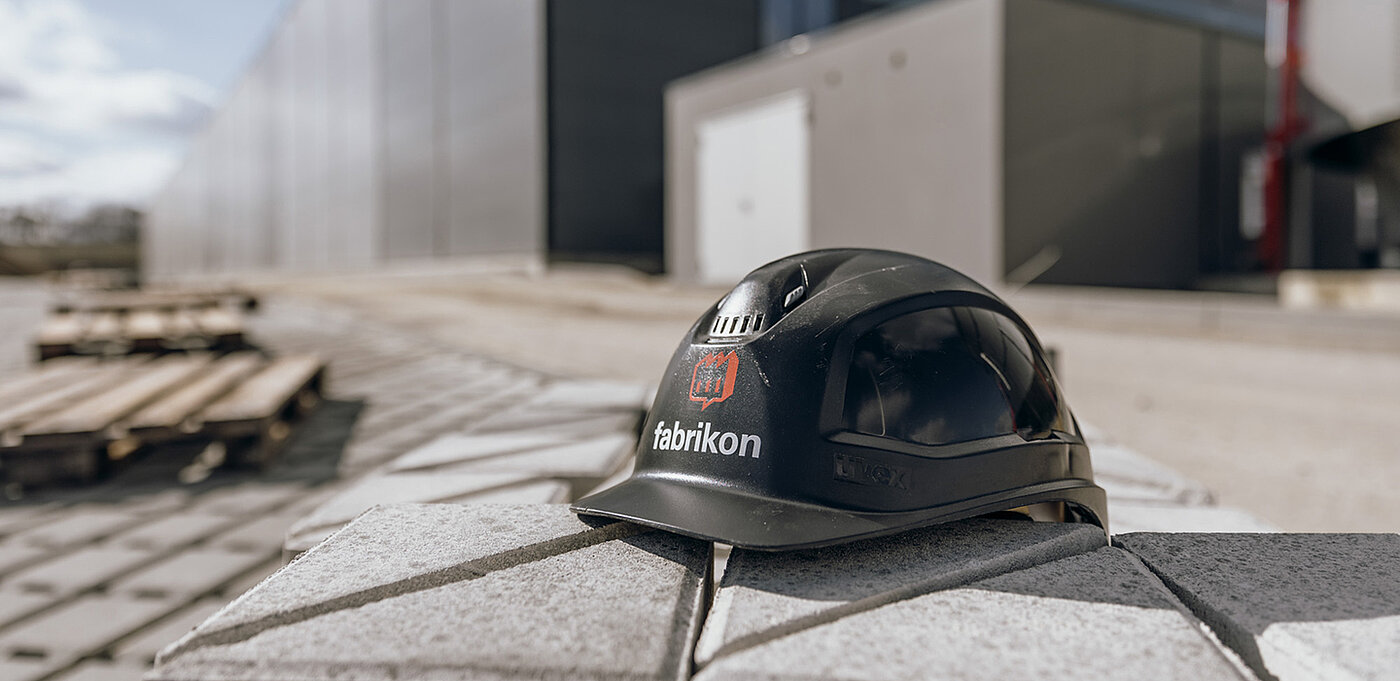 Schwarzer Schutzhelm mit dem Schriftzug "fabrikon" auf einer baustellenartigen Oberfläche.