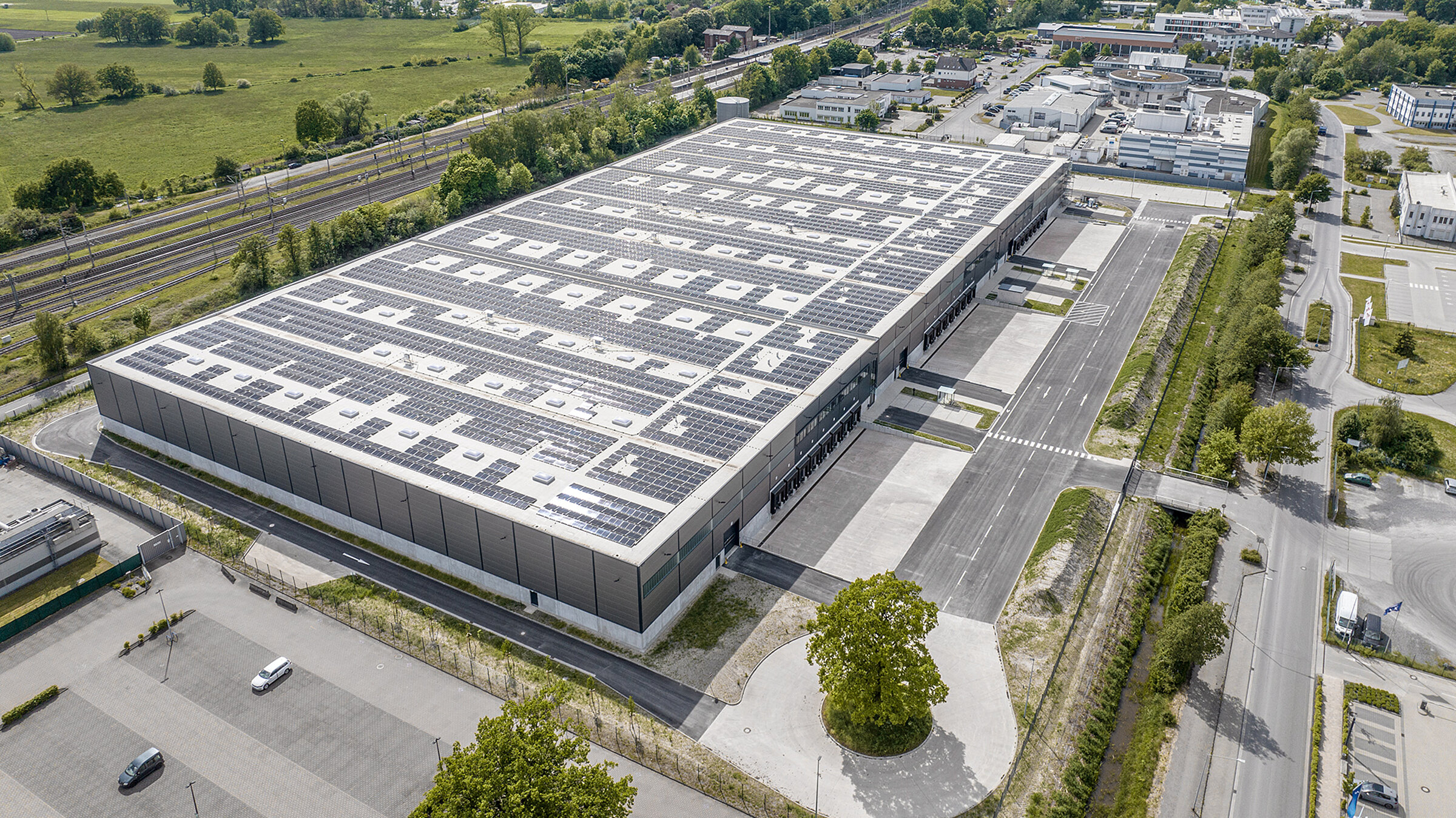 Luftaufnahme eines modernen Logistikgebäudes mit Solarpanelen und umliegenden Parkplätzen und Straßen.