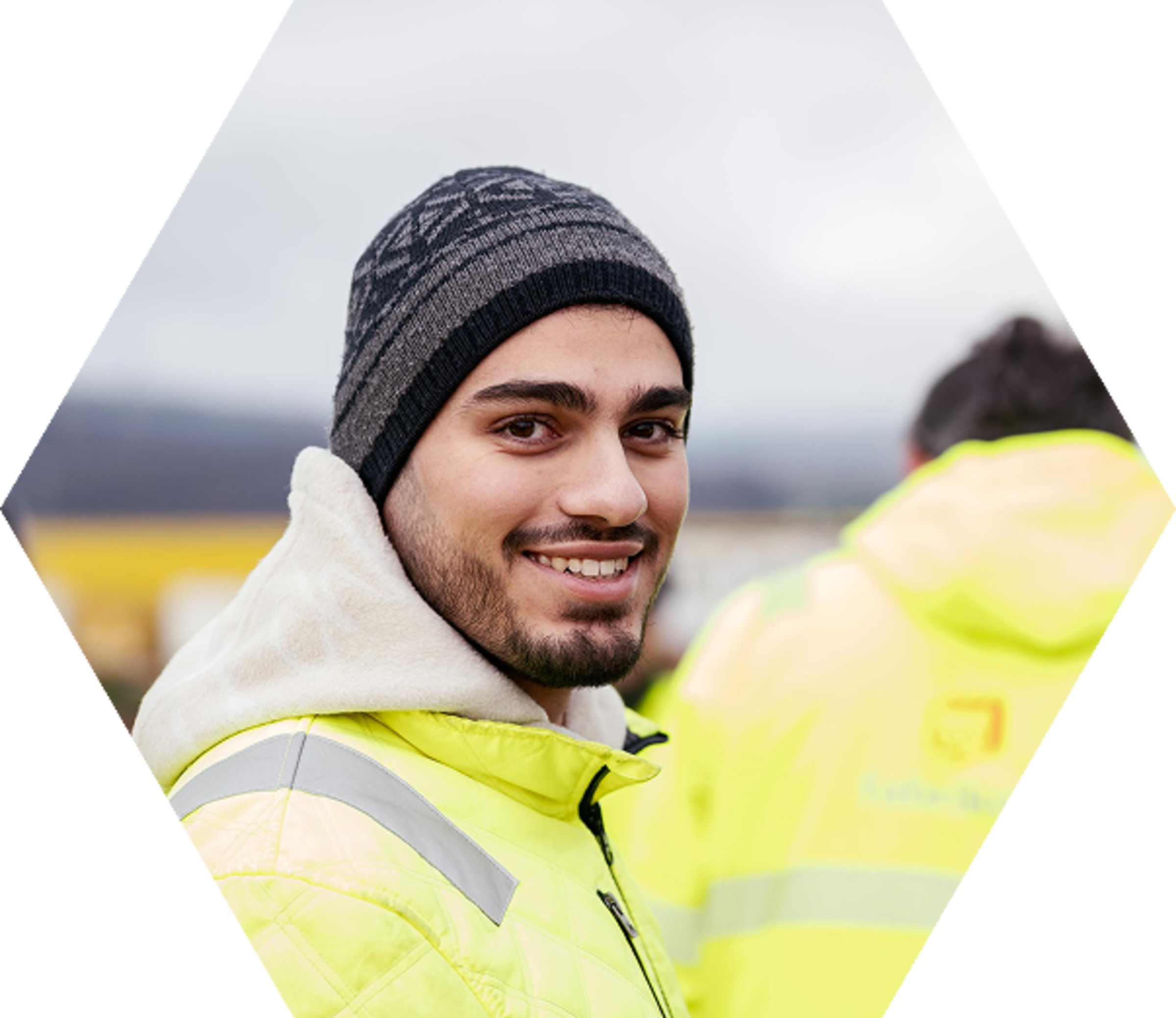 Lächelnder junger Mann in gelber Sicherheitsjacke und Wollmütze, mit verschwommenen Personen im Hintergrund.