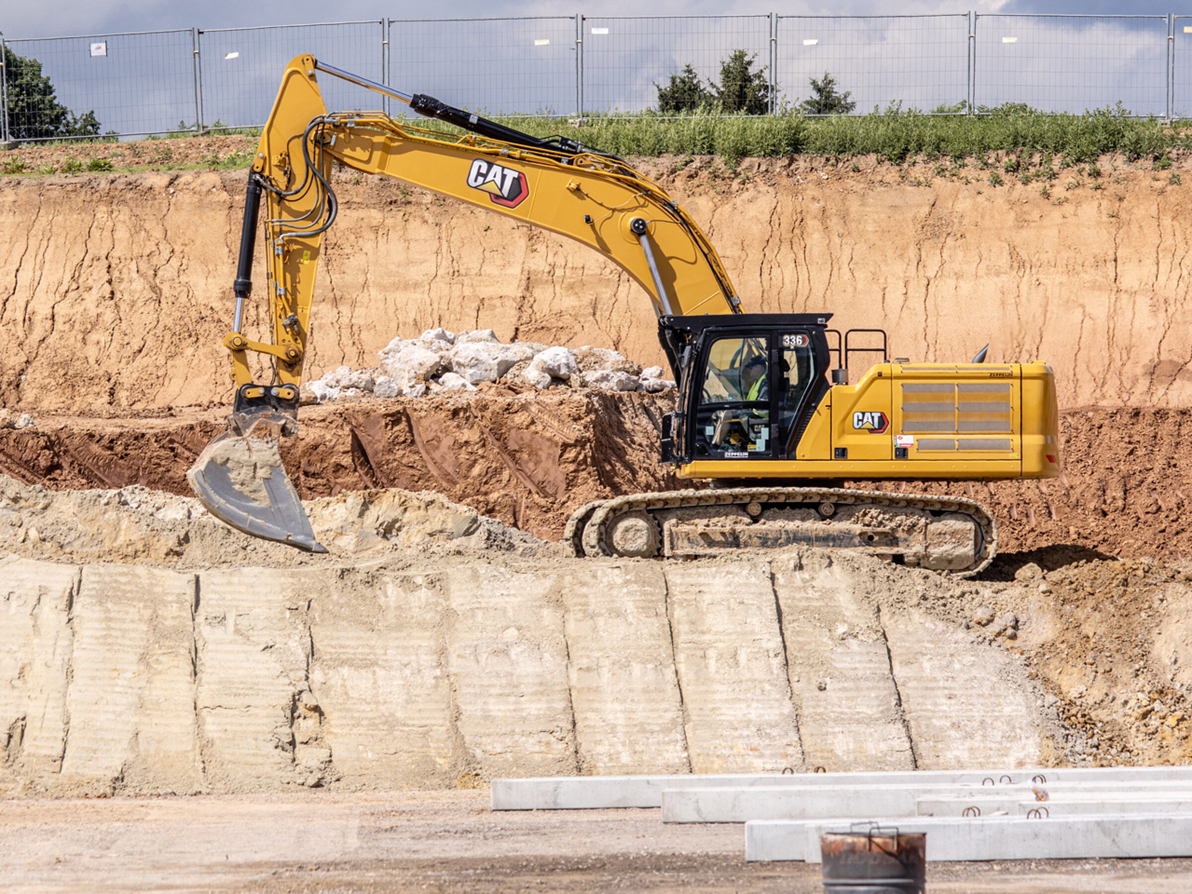 Excavator on a construction site lifts earth, with a cordoned-off area with a row of trees and construction fences in the background.