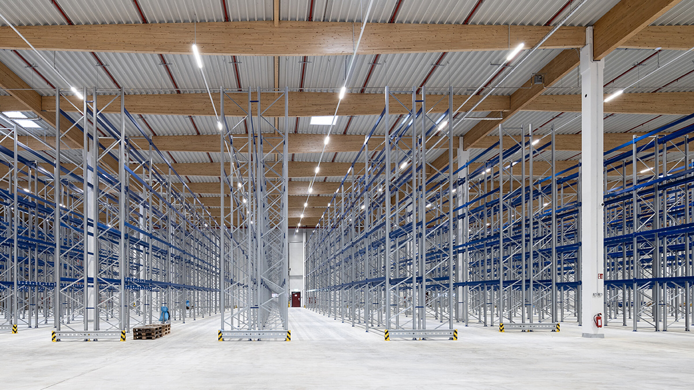 Leere Lagerhalle mit hohen Regalen auf der linken und rechten Seite, helle Beleuchtung und grauer Boden.