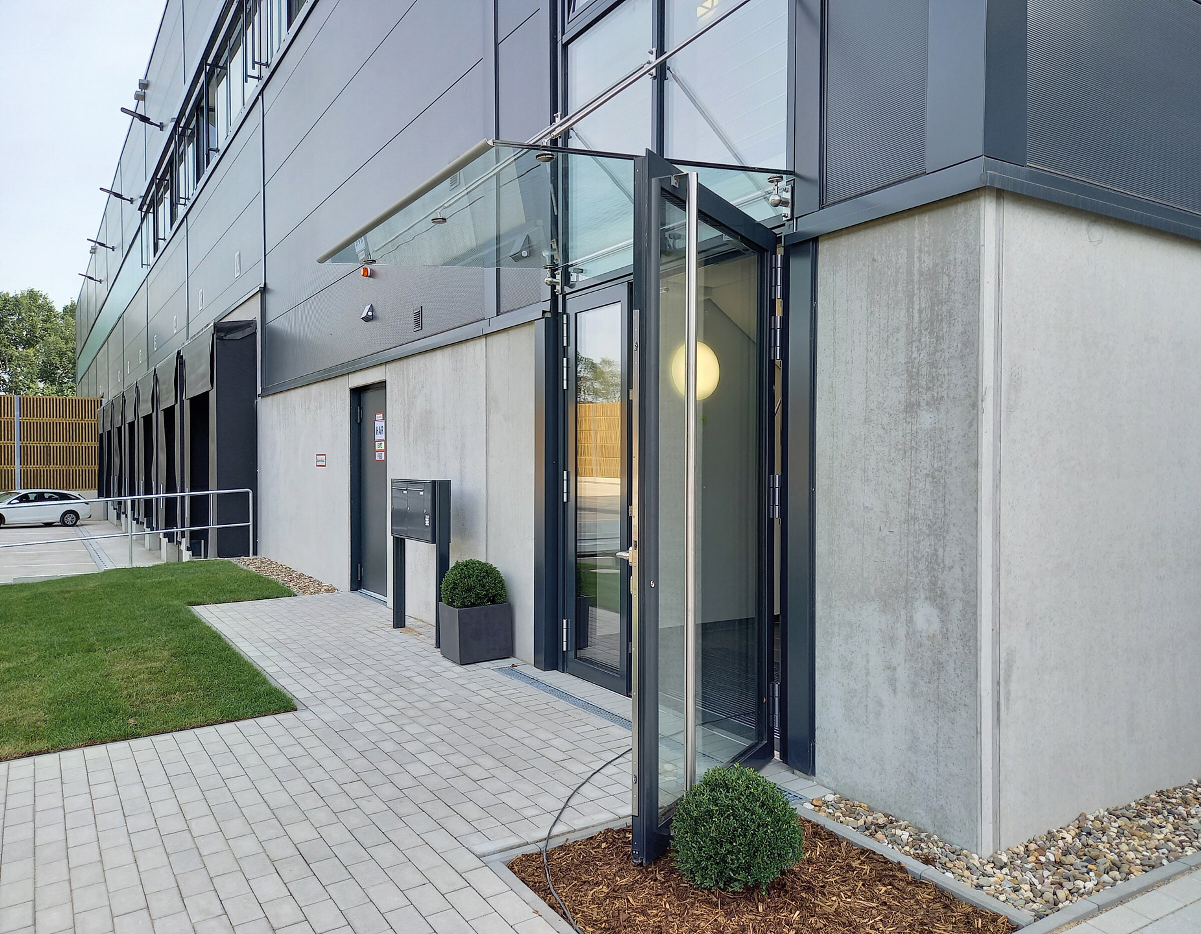 Eingang zu einem modernen Gebäude mit Glasdoppeltür, begrüntem Eingangsbereich und gepflastertem Weg.