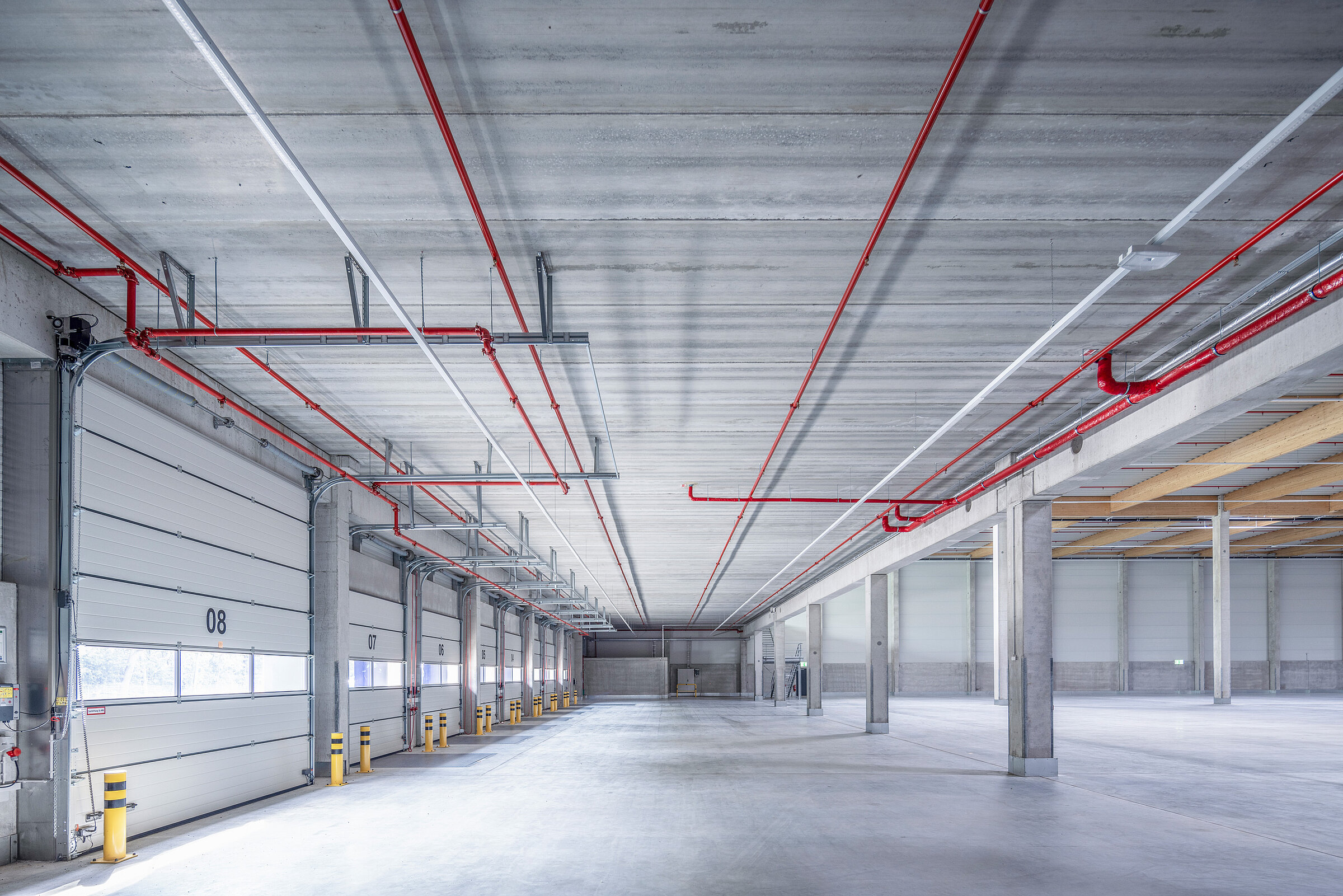 Interior view of a storage room with high ceilings, gray floor, and red and white pipes on the ceiling.