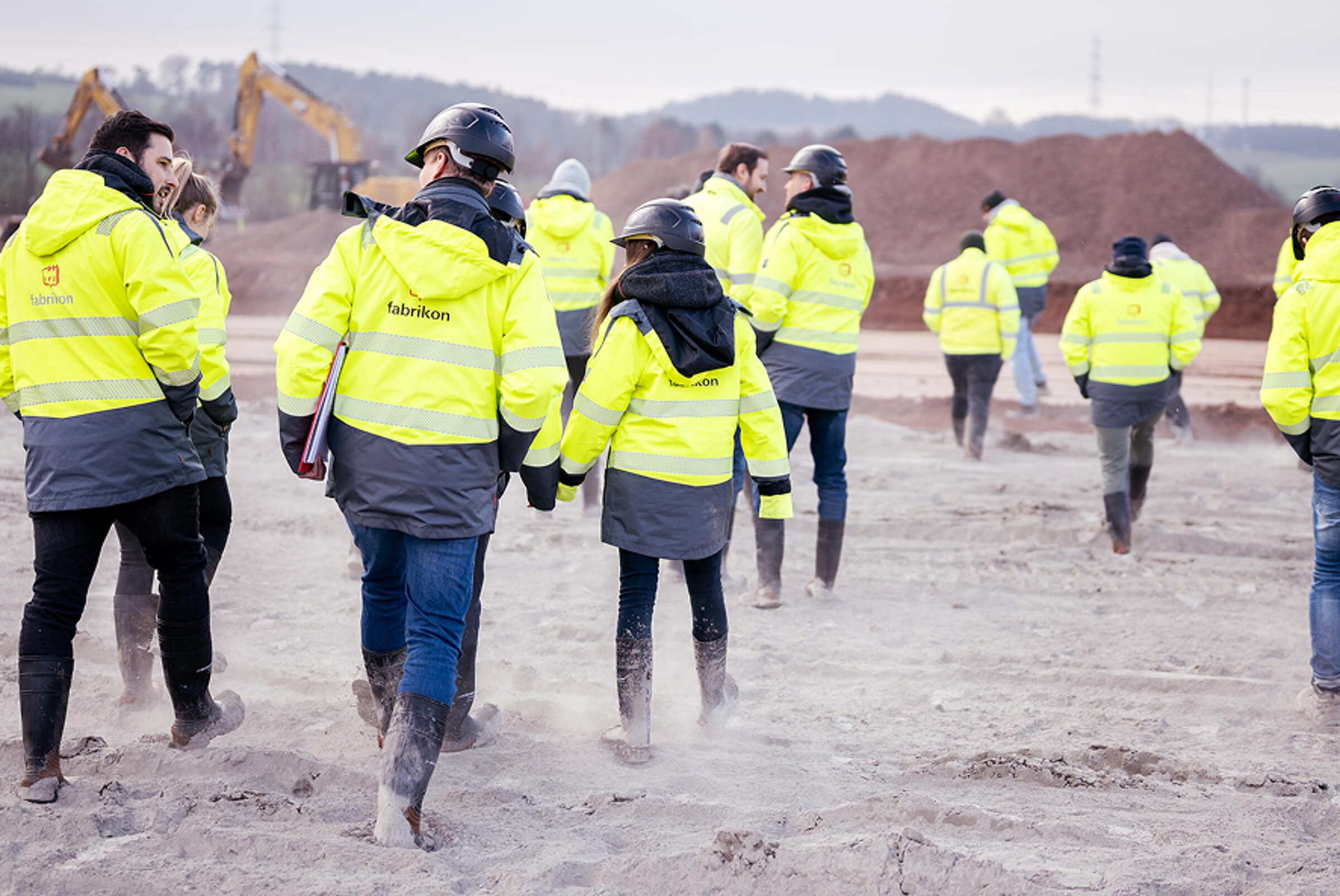 Grupp von Bauarbeitern in hellgelben Sicherheitsjacken und Helmen, die über eine Baustelle laufen.