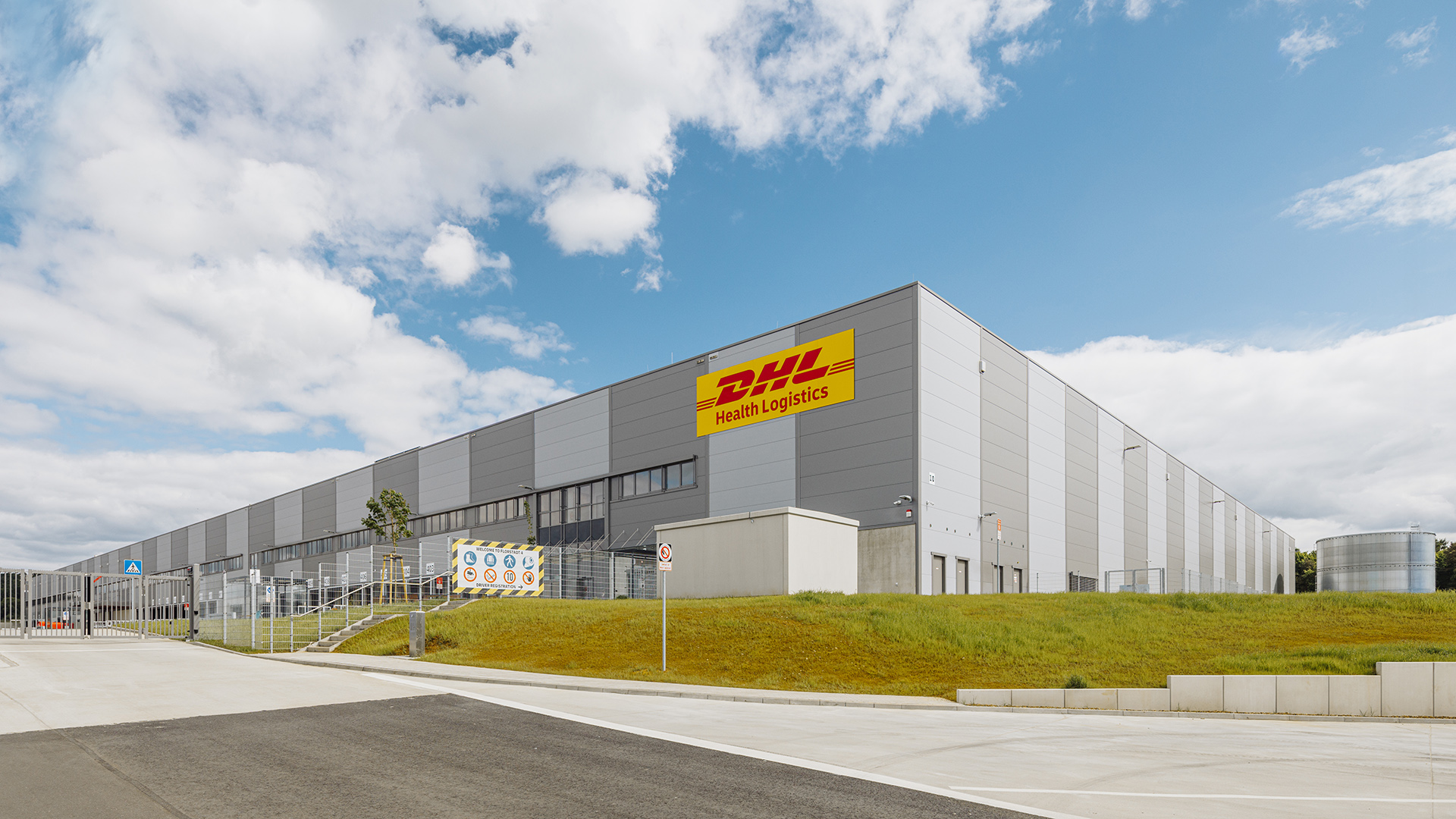 Großes, modernes Lagergebäude von DHL Health Logistics unter einem bewölkten Himmel.