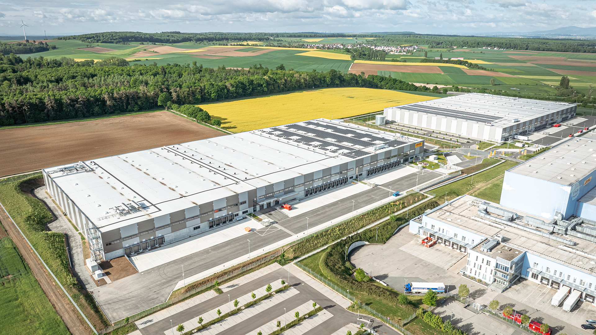 Luftaufnahme eines modernen Logistikgebäudes umgeben von grünen Feldern und landwirtschaftlichen Flächen.